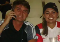 Bolsonaro e Michelle em foto antiga. Ele veste preto e segura um celular próximo ao ouvido. Ela usa um boné preto e veste uma camisa do Flamengo