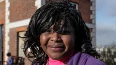 Uma foto do rosto da cientista. É uma mulher negra, com cabelos pretos. Ela está olhando para a foto e usa uma blusa rosa com um casaco roxo.