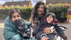 Um homem e uma mulher seguram uma criança pequena. O homem tem cabelo curto e escuro, barba e veste uma jaqueta acolchoada em tom verde-acinzentado. A mulher tem cabelo longo, escuro e ondulado, e usa um cachecol com estampa de oncinha. A criança tem cabelo escuro e veste um macacão acolchoado cinza-escuro com zíper