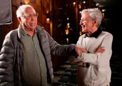 Os cantores Jards Macalé e Caetano Veloso dando risada, enquanto Macalé toca o braço de Caetano afetuosamente