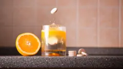 صورة لكوب مياه داخله قرص فوار وإلى يمينه المزيد من الأقراص الفوارة برتقالية اللون، وإلى يساره نصف برتقالة.