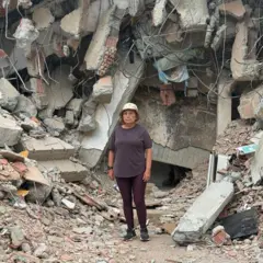 María Lourdes Pérez, vestida com calça e camisa marrom e gorro bege, em frente a uma montanha de escombros, cimento, pedras e partes de uma edificação