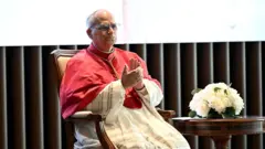 Папа Лев в белой сутане и красной моццетте сидит в кресле и хлопает в ладоши, на столике рядом с ним букет белых цветов