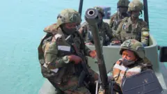 Abasirikare b'u Rwanda barimo babiri bagaragara ko ari abagore, umwe muri bo afite imbunda ya rutura, bari ku irondo mu bwato, mu mujyi uri ku cyambu wa Mocimboa da Praia, mu majyaruguru ya Mozambique, ku itariki ya 13 Kanama (8) mu 2021. Bambaye udupfukamunwa, impuzankano za gisirikare n'ingofero zikomeye za 'casques' za gisirikare, babiri muri bo bambaye amajaketi arinda kurohama mu mazi. 