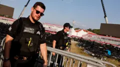 No primeiro plano um agente, vestindo uniforme preto e armado, ao lado de uma grade de proteção. No fundo, outro agente e mais ao fundo, cadeiras e uma arquibancada vazias.