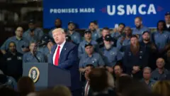 ダークスーツに赤いネクタイを着けたトランプ氏が、演台のマイクの前に立ち、話している。背後には、自動車業界の労働者らが並んで座っている