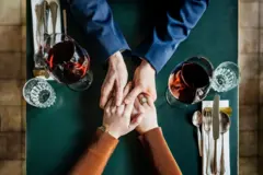 Pareja estrechando manos sobre una mesa con copas de vino.