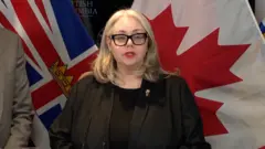 La ministra de Seguridad Pública y procuradora general de Columbia Británica, Nina Krieger, aparece con una chaqueta y una blusa negras frente a un telón con banderas de Canadá durante una rueda de prensa.