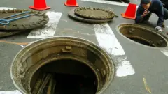 Otvorene šahte na putu i čovek koji baterijskom lampom ispituje jednu od njih