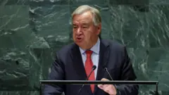 UN Secretary General Antonio Guterres dey give speech at di United Nations in New York. E dey stand against di distinctive green marbled backdrop at lectern, wearing red tie, dark suit jacket and blue shirt