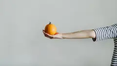 Braço esticado de uma pessoa de blusa listrada segura uma fruta na mão