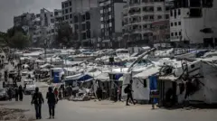 Pessoas passam por prédios destruídos e barracas improvisadas. As barracas são todas brancas e azuis. Alguns carros também podem ser vistos ao fundo.