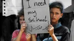 دو کودک دست‌نوشته‌ای را در دست دارند که روی آن نوشته شده است: «من به مدرسه‌ نیاز دارم.»