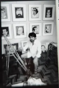 A imagem em preto e branco mostra Cláudio Seto, um homem barbudo e de óculos, agachado em um ateliê ou sala de trabalho, cercado por quadros com caricaturas pendurados na parede. À frente dele há um cavalete de madeira, enquanto outros retratos apoiados no chão e móveis aparecem ao redor. O ambiente tem piso de madeira em padrão geométrico e aspecto de arquivo fotográfico antigo.
