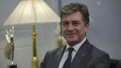 Gülümseyen bir adam, kameraya bakıyor. Arka planda ödül ve bir lamba var