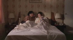 Un hombre (i) y una mujer conversan sonrientes en una cama.