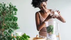 Stock photo shows a woman adding chia seends into a smoothie at home, there is a plant in the background.