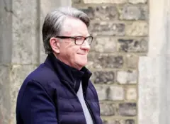 Peter Mandelson, con una camiseta gris, una chaqueta azul marino y gafas, sonríe mientras la policía se lo lleva tras su arresto a principios de esta semana.