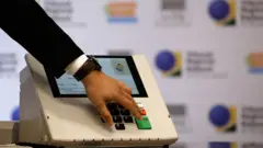 Close em urna eletrônica. Na frente dela aparece também uma mão apertando o botão de "confirma".
