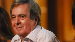 A imagem mostra Benedito Ruy Barbosa, de cabelos grisalhos, em primeiro plano, usando uma camisa listrada e olhando para a câmera com um leve sorriso. Ao fundo, desfocado e iluminado por luzes quentes, o ambiente sugere um estúdio ou palco, enquanto outras pessoas aparecem parcialmente nas laterais da cena.