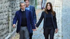 الرئيس الفرنسي السابق، نيكولا ساركوزي، مرتدياً بذلة رسمية ويسير في طريق إلى السجن، ويضع يده في يد زوجته كارلا بروني 