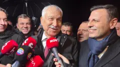 Karalar cezaevin çıkışında buruk bir gülümsemeyle açıklama yapıyor, önünde çeşitli televizyonların mikrofonları var, yanında CHP il başkanı özgür çelik var.