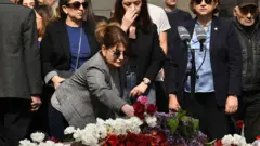 Bir kadın eğilmiş karanfillerin üzerine çiçek bırakıyor. 
