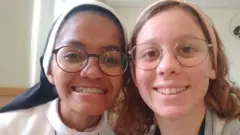 Fotografia colorida mostra duas mulheres de óculos sorrindo para a foto. A da esquerda usa um hábito de freira e a da direita usa uma passadeira rosa no cabelo