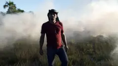 Homem com chapéu e camisa vermelha posa com fumaça e vegetação de cerrado ao fungo.