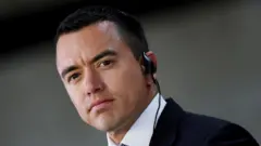 Foto de Noboa, serio, con un auricular y vestido con un traje oscuro.