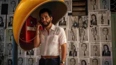 Wagner Moura aparece no papel de um professor em O Agente Secreto. O ator fala em um telefone público vermelho, sob uma cabine amarela, com expressão séria e concentrada. Ao fundo, cartazes políticos antigos cobrem a parede, sugerindo um contexto de tensão política e ambientação histórica.