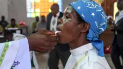 The white embroidered sleeve of a Catholic cleric in Kenya placing a communion wafer of the tongue of a kneeling worshipper wearing white robes and a blue headscarf
