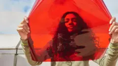 Bir kadın kameraya bakarken yüzüne ve omuzlarına bir kırmızı filtre tutuyor. Arka planda gökyüzü görünüyor. 