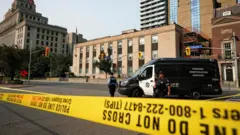 3階建ての建物の前の通りに、黄色い規制線が張られている。路上には黒と白の警察車両と、警官2人が見える
