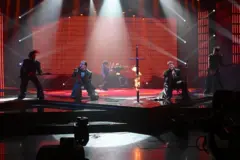 muškarci klečeći pevaju na sceni, obučeni su u crno, između njih je mač koji gori 