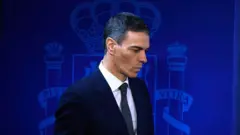 İspanya Başbakanı Pedro Sanchez, lacivert zemin üzerinde İspanya armasının yer aldığı bir arka plan önünde takım elbise ve kravatla görülüyor.