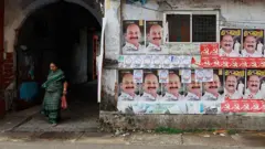 Uma mulher sai de uma colônia residencial enquanto cartazes da campanha eleitoral do candidato da Frente Democrática Unida (UDF) Mohammed Shiyaz e do candidato da Frente Democrática de Esquerda (LDF) K J Maxy são exibidos ao longo de uma estrada antes da eleição para a Assembleia em Kochi, Índia, em 4 de abril de 2026.