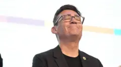 白い背景の檀上に黒い上着と丸襟のシャツ姿で立った、眼鏡をかけたバーナム氏が、両目を閉じて口をぎゅっと結んで微笑み、上を向いている