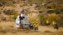 Yerde hareketsiz duran beyaz bir helikopterin yanında dört itfaiye çalılık bir alanda görünüyor. 