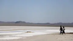 Un hombre y una mujer caminan por el lecho seco del lago salado Hoz-e Soltan en Qom: se puede ver sal en la superficie y montañas en la distancia.