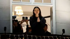 María Corina Machado en el balcón de un hotel en Oslo, Noruega. Está de pie, cabello negro hasta los hombros y sonríe mientras junta las palmas de las manos. Viste un abrigo negro. 