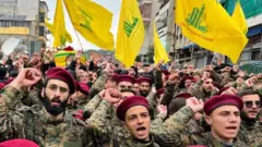 Des membres du Hezbollah en uniforme militaire défilent avec les drapeaux du groupe en arrière-plan.