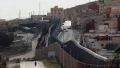 صورة التُقطت من الجانب الإسباني في 24 فبراير/شباط 2024 تُظهر الجدار والسياج اللذين يُحددان الحدود بين المغرب (يمين) وجيب مليلية الإسباني في شمال أفريقيا (يسار)