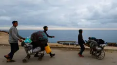 Três homens empurram um carrinho de supermercado carregado de pertences pessoais. O mar pode ser visto ao fundo
