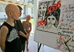 Um grupo de crianças, duas delas carecas, olha para cartazes com os dizeres "1986, 2006".