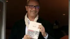 Kamel Daoud, avec des lunettes à monture noire, sourit alors qu'il se tient devant une fenêtre ouverte, tenant un exemplaire blanc de son livre primé. Il porte un blazer sombre et une chemise blanche, ainsi qu'une écharpe de couleur crème légèrement enroulée autour du cou.