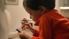 Um garoto com uma camisa vermelha de manga comprida está concentrado em um smartphone vermelho enquanto está sentado em um ambiente interno. O fundo pouco iluminado acrescenta uma atmosfera aconchegante.