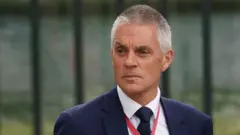 A close crop of Tim Davie shows his head and shoulders as he frowns looking away from camera while wearing a suit and tie and a red lanyard around his neck, pictured outside Westminster Abbey ahead of King Charles's coronation in May 2023.