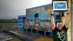 Imagem mostra diretor segurando placa de prêmio da escola. Ao fundo está o prédio da escola