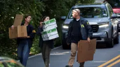 Três agentes do FBI em uma rua carregam caixas e papelões.